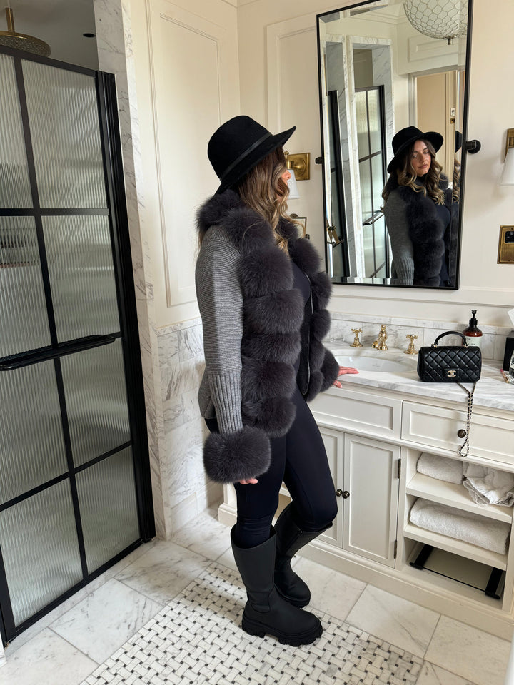 Frau in einem VELORIA – Winter Cardigan mit Kunstpelzkragen, kombiniert mit modischer Winterkleidung und Hut.