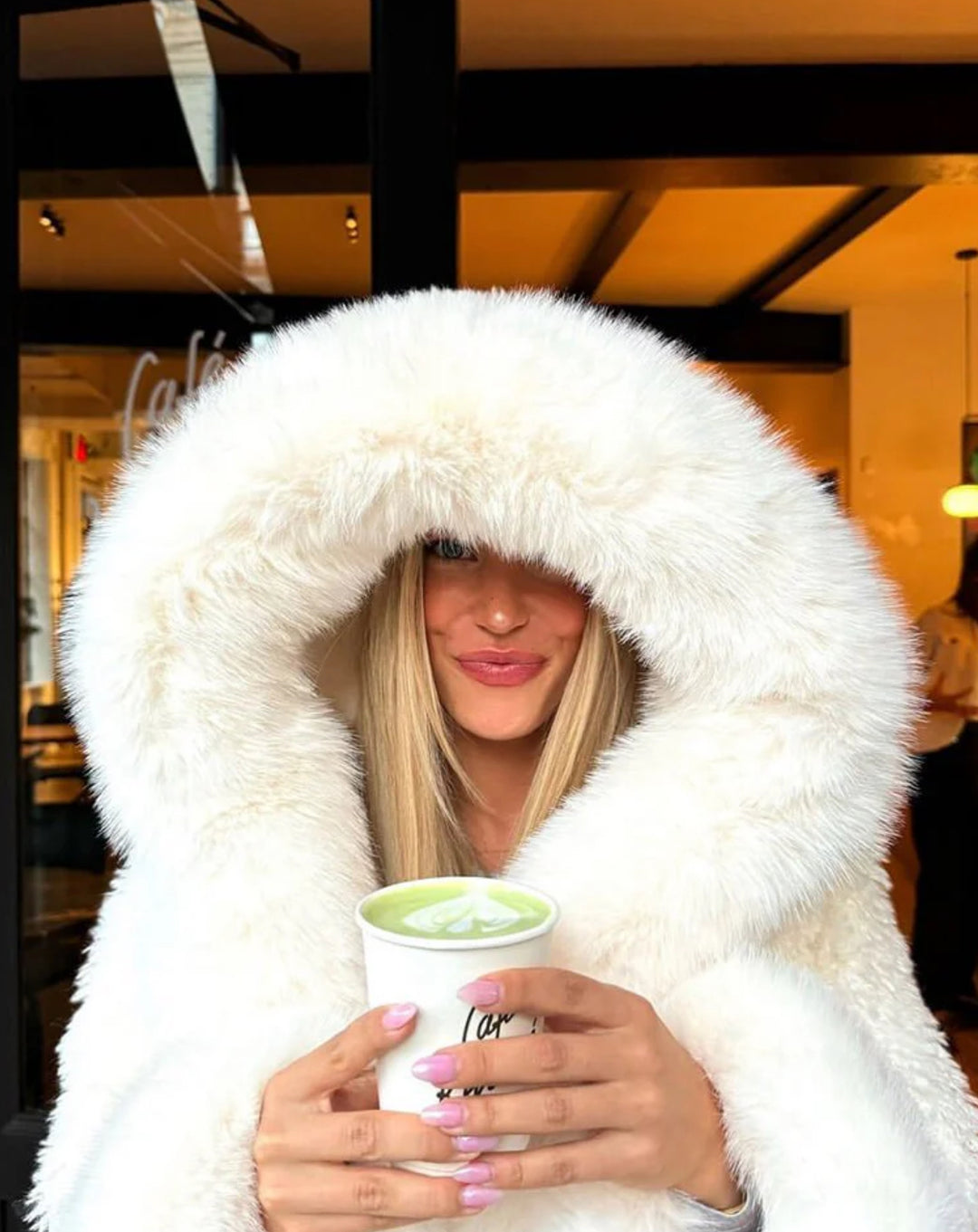 AMÉLIE  – Flauschige Felljacke mit Kapuze, die eine Frau mit einem Getränk in einem Café trägt.
