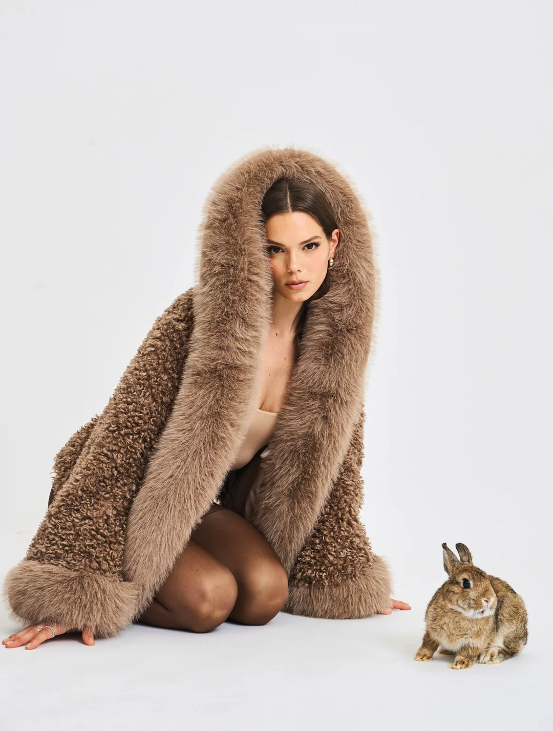 AMÉLIE  – Flauschige Felljacke mit Kapuze, getragen von einem Model mit einem Kaninchen auf dem Boden.