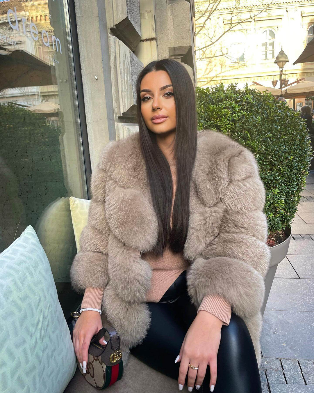 Frau trägt SOPHIE - eleganter Kunstpelzmantel von Miraee Paris mit modischer Handtasche in einem Café.